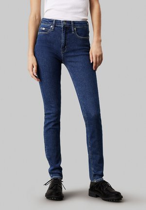 Dark blue skinny jeans with a mid-rise waist, front pockets, and subtle stitching details. Paired with black lace-up shoes.