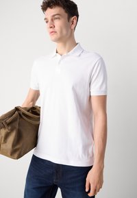 Hvit polo skjorte med korte ermer og en subtil logo; brukt sammen med mørkeblå denim, holder en brun lærveske. Enkelt og rent design.