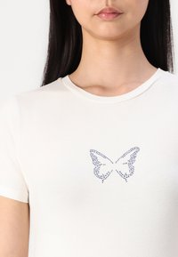 T-shirt crème en tissu doux avec un design de papillon créé avec des rhinestones bleus sur la zone de la poitrine.