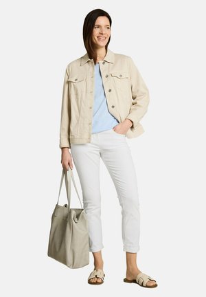 Kobieta w beżowej kurtce, jasnoniebieskiej koszuli, białych spodniach, beżowych sandałach, trzyma dużą beżową torebkę, stoi i uśmiecha się na białym tle.