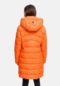 Mujer con un abrigo largo de color naranja brillante y acolchado con capucha, vista desde atrás sobre un fondo claro y sencillo.