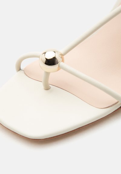 Close-up van een witte sandaal met open teen en dunne bandjes met een glanzend gouden kraal detail bij het teengebied op een beige binnenzool.