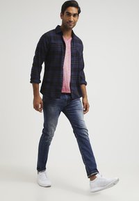 Camisa a cuadros en tonos azul marino y púrpura sobre una camiseta de cuello en V rosa claro, combinada con jeans oscuros de corte ajustado y zapatillas blancas.