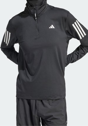 Osoba nosząca czarny, długi rękaw top do biegania Adidas z białymi logo z trzema paskami na rękawach i piersi, w połączeniu z czarnymi szortami.