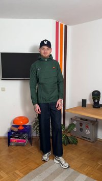 Green windbreaker with a hood, front pocket, and logo. Paired with dark denim pants and gray sneakers. Simple design with practical features.