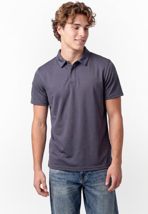 Jeune homme aux cheveux bouclés portant un polo gris foncé et un jean bleu, regardant vers le bas et souriant légèrement sur un fond blanc.