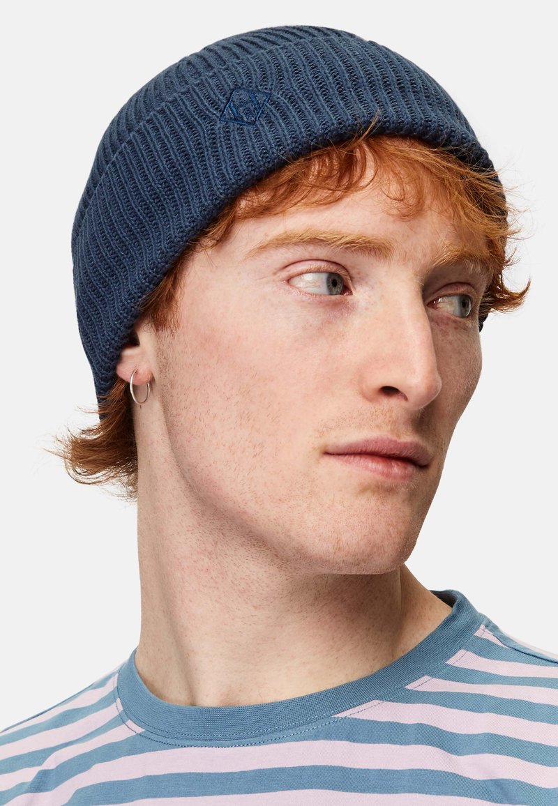 Mörkblå stickad mössa med ribbad textur och logodetalj. Modellen bär en randig skjorta i blått och rosa.