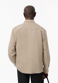 Brązowa i kremowa koszula w kratę z kołnierzykiem, długimi rękawami i mankietami na guziki; posiada prosty dół i detale na plecach.