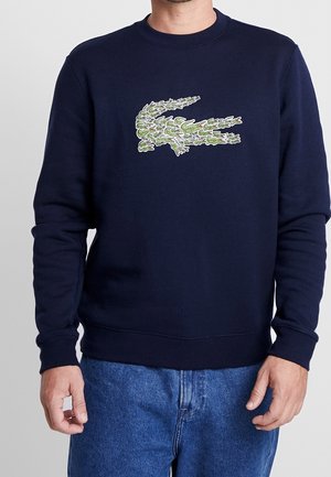 Mężczyzna w granatowej bluzie z zielonym logo krokodyla i niebieskich dżinsach, stojący na tle jasnego, jednolitego tła.