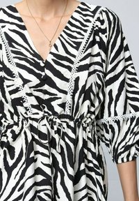 Robe noire et blanche à motif zèbre avec un décolleté en V profond, taille élastique et manches bouffantes. Elle présente des détails en dentelle et des boutons.