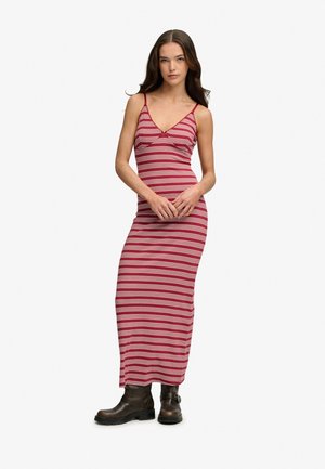 Kobieta w czerwoną i białą sukienkę maxi bez rękawów w paski, zestawioną z czarnymi botkami do kostki, stojąca na jednolitym jasnym tle.