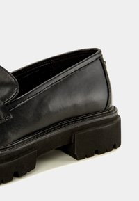 Mocassin en cuir noir présentant une texture lisse, un profil bas et une semelle en caoutchouc épaisse avec une bande de roulement à motif pour une meilleure adhérence.