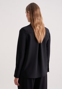 Camicia nera a maniche lunghe con texture liscia, polsino con bottoni e orlo leggermente curvato. Presenta un colletto classico e un design minimal.