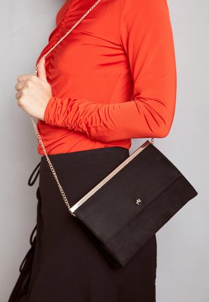 Person iført en rød bluse med lange ærmer og sort nederdel, der holder en sort clutch med en guldkæde.