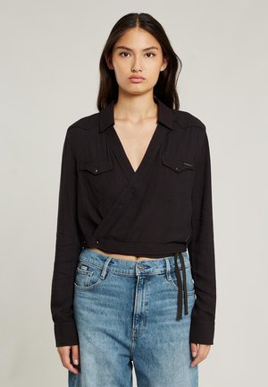 Czarna koszula z długim rękawem, kołnierzykiem i kieszeniami z przodu. Posiada cropped design i zawiązanie w talii. Noszona z jeansami o wysokim stanie.