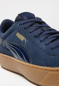 Gros plan d'une basket Puma bleu marine avec dessus en daim, bande logo bleu foncé, texte Puma doré et semelle en caoutchouc beige.