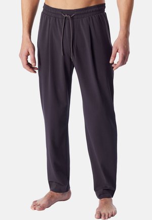Mann trägt dunkelgraue elastische Bundhose mit Kordelzug, steht barfuß vor hellem Hintergrund, Oberkörper von der Taille abwärts sichtbar.