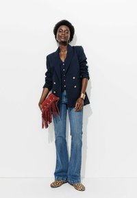 Blazer bleu marine croisé, gilet noir, jeans en denim évasés, pochette frangée rouge et ballerines à imprimé léopard. Les accessoires comprennent un bracelet manchette en or.