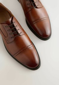 Barna bőrdressz-cipők sima felülettel, lekerekített orral és minimális varrási részletekkel. Laposságú fűzőkkel és texturált talppal rendelkezik.