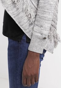 Grå stickad cardigan med fransade kanter och ribbade ärmmuddar, bärs över en svart topp och matchas med blå jeans.