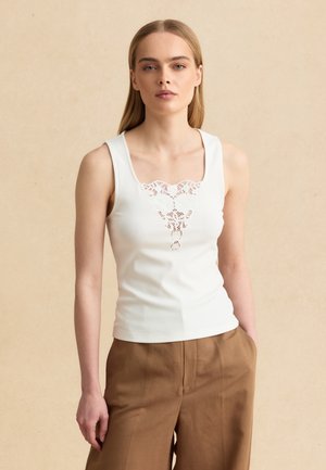 Woman wearing a white sleeveless top with lace detail at the neckline and high-waisted brown pants, standing against a beige background.