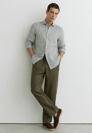 Uomo appoggiato a una parete bianca che indossa una camicia a righe grigio chiaro con bottoni, pantaloni verde oliva e scarpe scure senza lacci.
