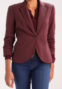 Femme portant un blazer ajusté marron foncé sur un haut assorti et un jean bleu, debout les bras détendus le long du corps.
