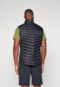 Black puffer vest with quilted design, high collar, and side panel accents. Worn over a dark green short-sleeve shirt and black shorts.