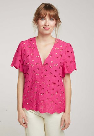 Chemisier rose avec un design à œillets floraux, manches courtes flutter, et un décolleté en V profond. Ourlet festonné avec des accents de boutons. Tissu léger.