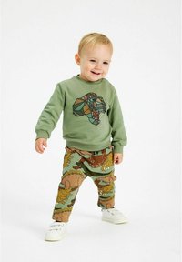 Zeleni dukserica s geometrijskim printom dinosaura, uparen s hlačama u zeleno-smeškastom uzorku s crtanih životinjama, i belim cipelama.