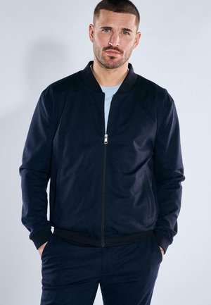 Man with short hair and beard wearing a zipped navy blue jacket and matching pants, standing with hands in pockets against plain background.