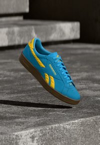 Modro-žuti Reebok čevelj z rjavim podplatom, ki lebdi nad teksturirano betonsko površino in stopnicami v mehki svetlobi.