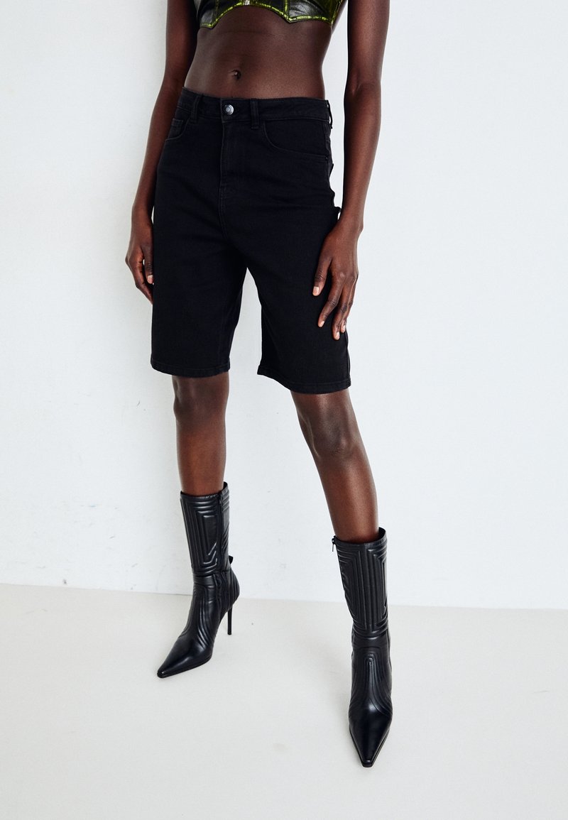 Černé džíny šortky s klasickým střihem, které mají pět kapes a zapínání na knoflík. V kombinaci s texturovanými černými vysokými podpatky.