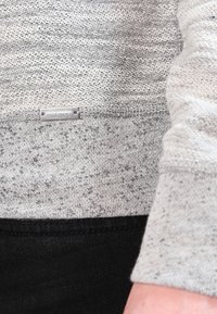 Suéter gris texturizado con un dobladillo negro, que presenta una pequeña etiqueta de logo plateado. La tela muestra una mezcla de fibras oscuras y claras.