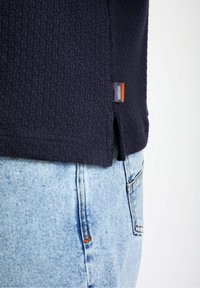 Marineblå tekstureret top med sideslids, parret med lysblå denim, som har et lille mærketag ved sømmen.