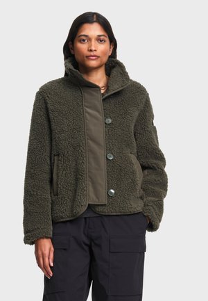 Femme portant une veste polaire texturée verte avec de gros boutons et un pantalon cargo foncé, debout devant un fond clair uni.