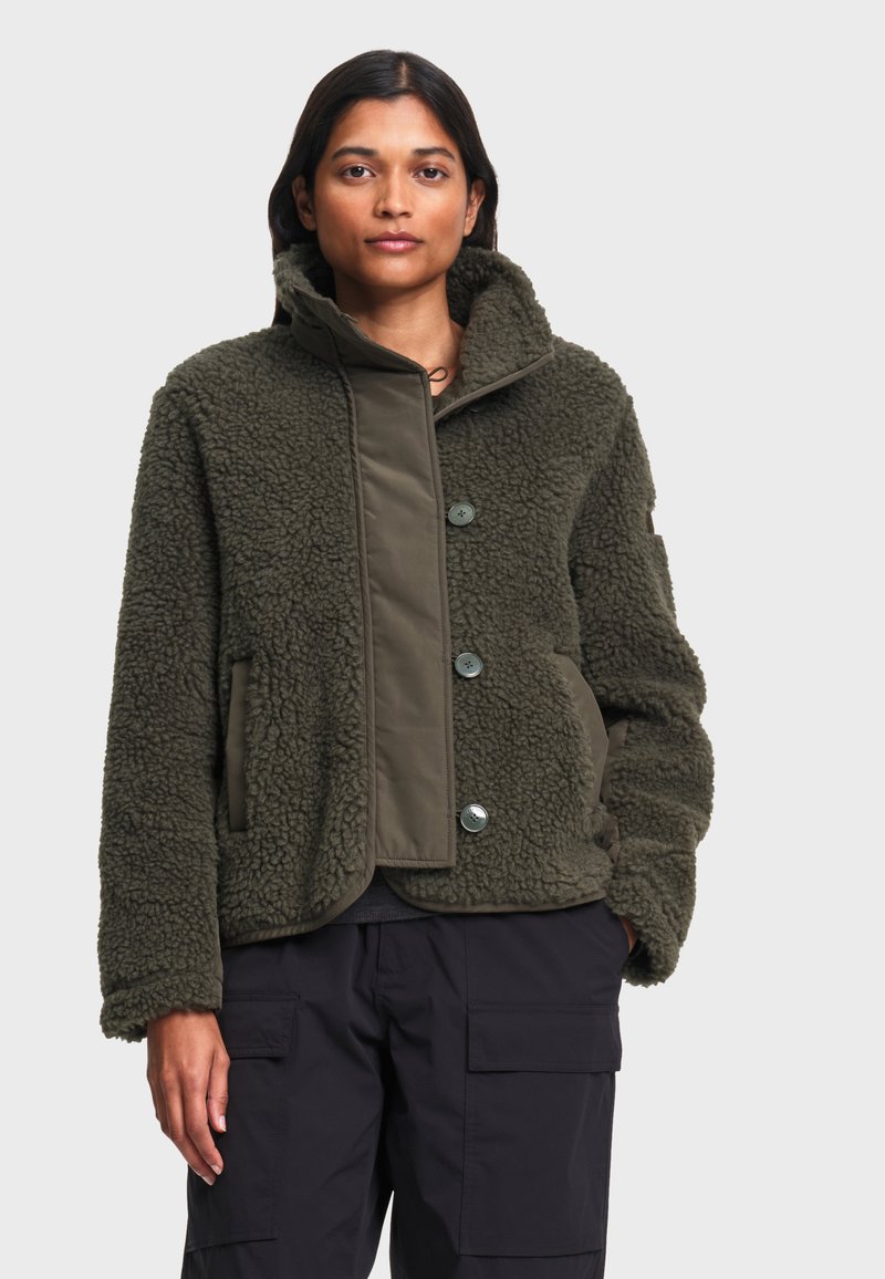 Femme portant une veste polaire texturée verte avec de gros boutons et un pantalon cargo foncé, debout devant un fond clair uni.