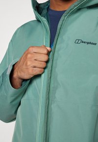 Chaqueta impermeable verde con cremallera completa, capucha ajustable y detalle de logo. La textura suave y el diseño sin costuras realzan la funcionalidad.