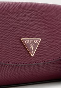 Bordeaux gekleed portemonnee met een gouden driehoekig logo met "GUESS" en een vraagteken. Verfraaid met stiksels langs de randen.