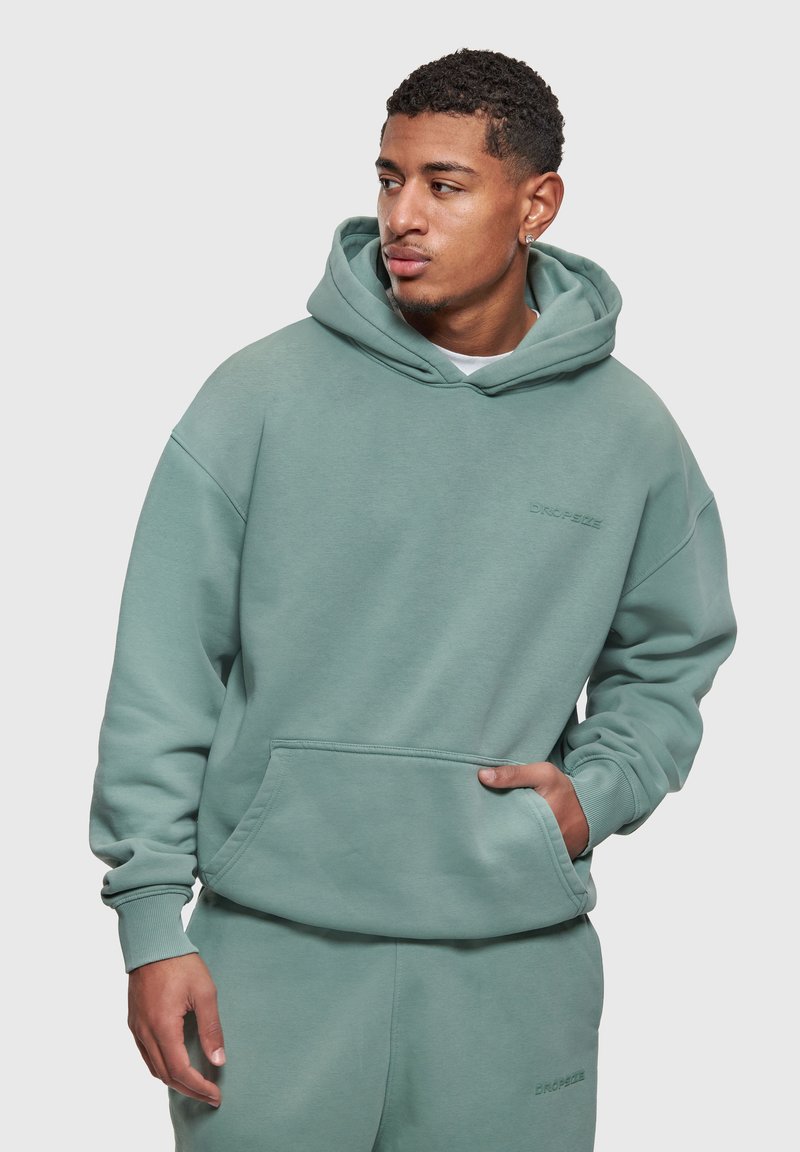 Hombre con sudadera con capucha verde y pantalones deportivos a juego, con la mano en el bolsillo, mirando hacia un lado contra un fondo claro y liso.