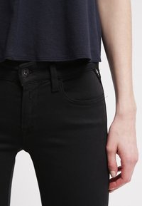Vaqueros de mezclilla negros de corte slim, con cierre de botón en la parte delantera, cinco bolsillos y una textura suave. Combinados con una camiseta corta de color azul marino.