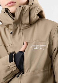 Beige Outdoor-Jacke mit hohem Kragen, Reißverschluss und gesticktem Logo. Verfügt über verstellbare Bündchen und mehrere Taschen.