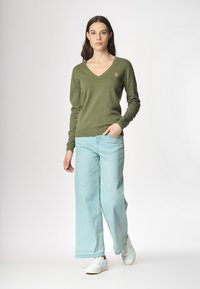 Modello in posa con un maglione verde a V e jeans a gamba larga blu chiaro, indossando sneaker bianche, su uno sfondo neutro.