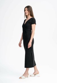 Schwarzes, knielanges Wickelkleid mit kurzen Ärmeln, seitlichem Schlitz und Kordelzug in der Taille. Das Modell trägt weiße Blockabsatzsandalen.