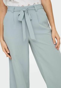Pantalon bleu clair ajusté avec une coupe à jambes larges, doté d'une ceinture en tissu nouée, de plis à l'avant et d'une finition lisse et texturée.