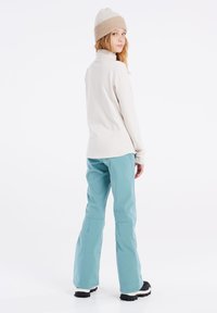 Aangemeten beige langemouwen top gecombineerd met losse turquoise broeken, gedragen met een beige en lichtbruine gebreide beanie en zwarte laarzen.