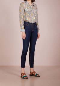 Camisa de estampado floral con detalles en dorado, jeans ajustados de color azul oscuro y sandalias marrones con acentos en verde azulado. Texturas suaves y diseño entallado.