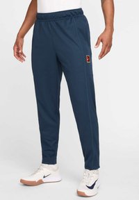Marinblå sportiga joggers med elastisk midja, sidofickor och en flerfärgad patch-logotyp. Tillverkade av mjukt, texturerat tyg.