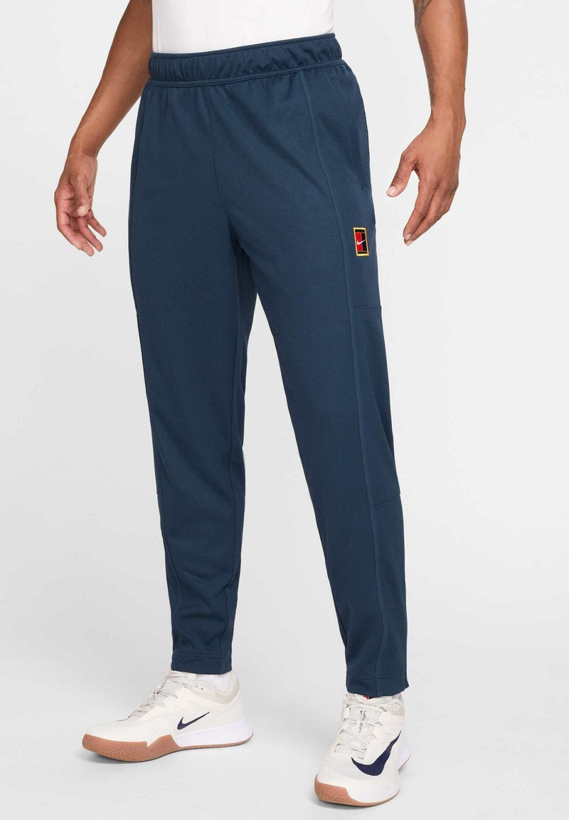 Marinblå sportiga joggers med elastisk midja, sidofickor och en flerfärgad patch-logotyp. Tillverkade av mjukt, texturerat tyg.
