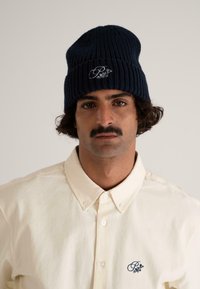 Navy ribbed knit beanie with embroidered design; cream button-up shirt with collar and small navy logo on the left chest.